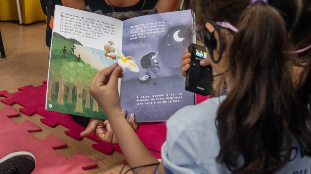 Criança estudante do Centro de Ensino Especial de Deficientes Visuais (CEEDV) escutando a audiodescrição do livro Pepita, uma abelhinha sem ferrão.